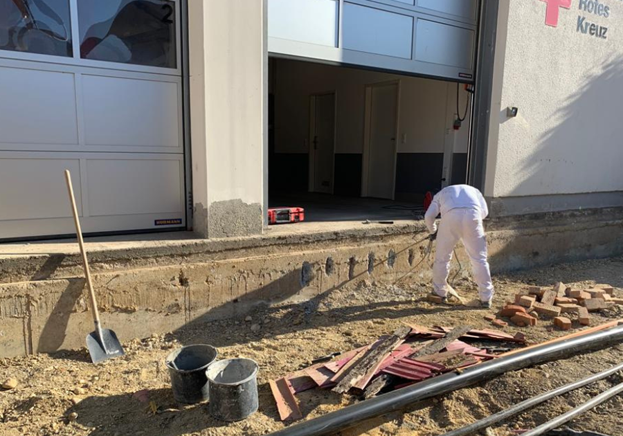 Handwerker bereitet den Betonsockel vor; freigelegte Kante und Ankerpunkte sichtbar.