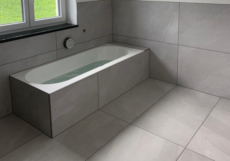 Moderne Badewanne mit flächenbündiger Verkleidung aus hellgrauen, großformatigen Feinsteinzeugfliesen. Die Wanne ist vor einem Fenster in eine geflieste Vorwand integriert, der Boden ist ebenfalls durchgängig mit den gleichen Fliesen gestaltet. Die Wanne ist halbvoll mit Wasser.
