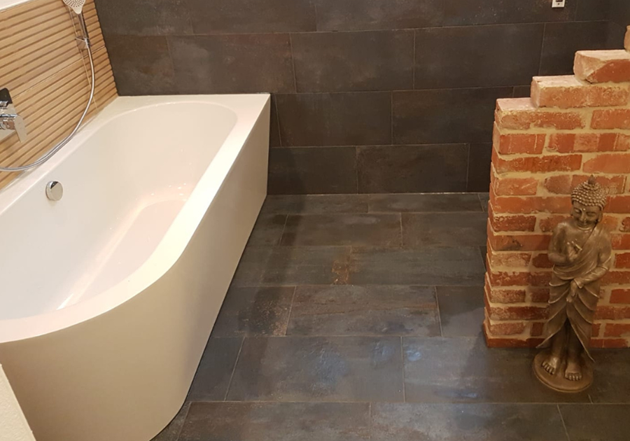 Das Bild zeigt eine weiße Badewanne, die an einer Wand mit horizontal strukturierten Fliesen steht. Der Boden ist mit großen, dunkelgrauen Fliesen ausgelegt, die auch an der Wand hinter der Wanne weitergeführt werden. Rechts im Bild befindet sich eine kleine Mauer aus roten Ziegelsteinen, vor der eine dekorative Figur steht.