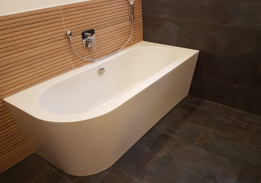 Eine weiße Badewanne steht an einer Wand mit hellbraunen, horizontal strukturierten Fliesen. Darüber befindet sich eine Armatur mit Handbrause. Der Boden ist mit großen, dunkelgrauen Fliesen ausgelegt, und die rechte Wand besteht aus denselben dunklen Fliesen.