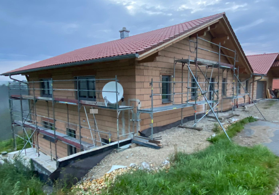Ein Haus im Bau mit unverputzter Ziegelfassade, Gerüsten und sichtbarer Sockelabdichtung. Vor dem Gebäude liegen Kies und Erdreich, im Vordergrund wächst Gras.