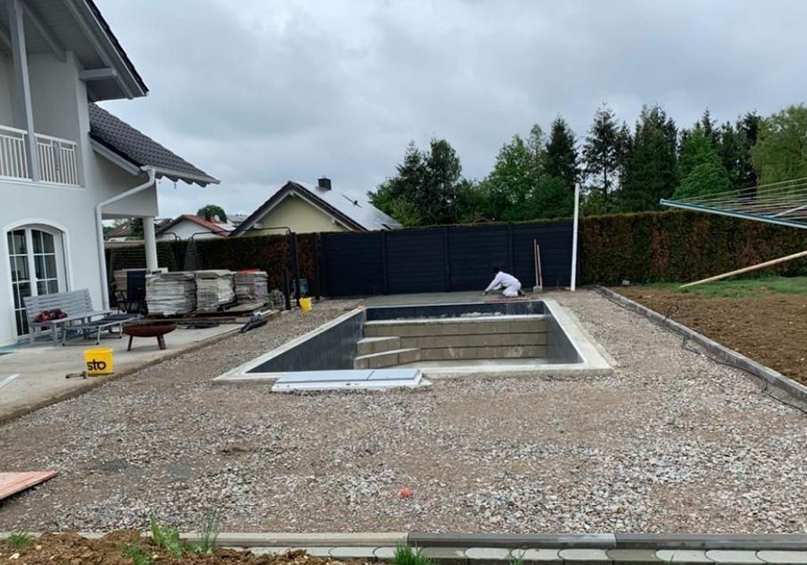 Ein Garten mit einem im Bau befindlichen rechteckigen Pool, umgeben von Schotter und Baumaterial. Links steht ein Haus mit Terrasse, auf der Bauutensilien und gestapelte Fliesen liegen.