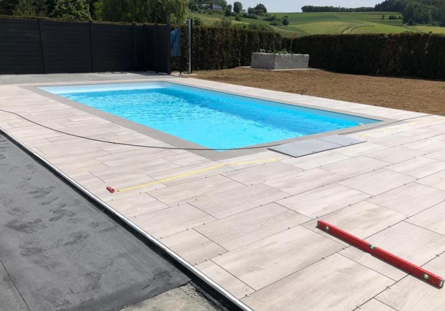 Ein rechteckiger Außenpool mit hellblauem Wasser ist von großen, hellen Steinfliesen umgeben. Im Vordergrund liegen Werkzeuge, was auf laufende Bau- oder Verlegearbeiten hinweist. Im Hintergrund befinden sich eine Hecke, eine schwarze Zaunanlage und grüne Wiesenlandschaft.