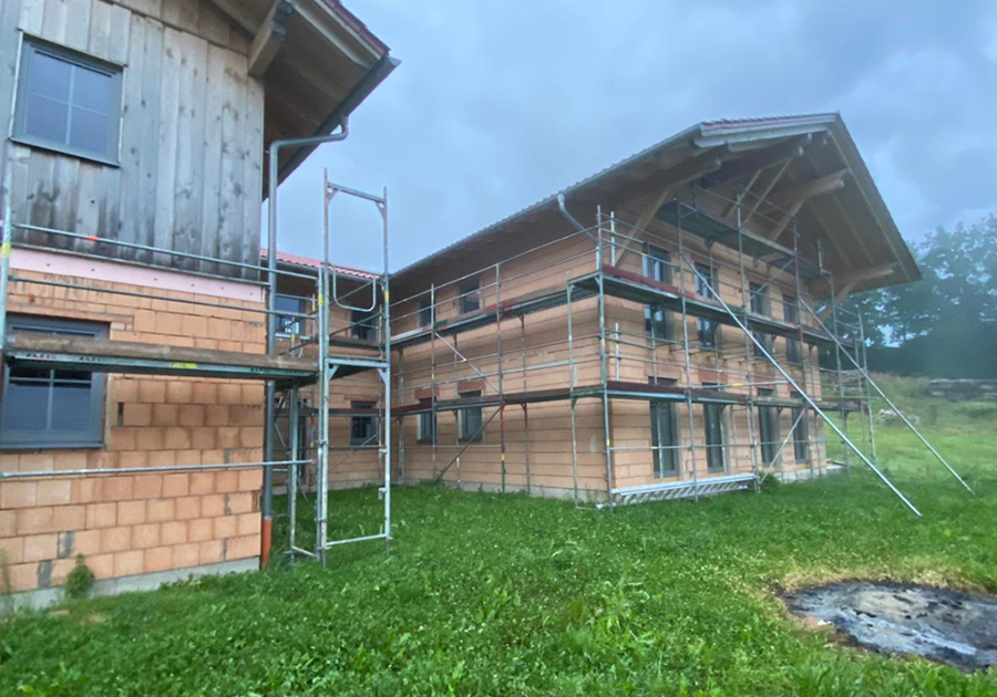 Ein Haus im Bau mit Gerüsten an den Außenwänden. Die Fassade besteht aus unverputzten Ziegeln und Holzelementen, und das Gebäude steht auf einer grasbewachsenen Fläche.