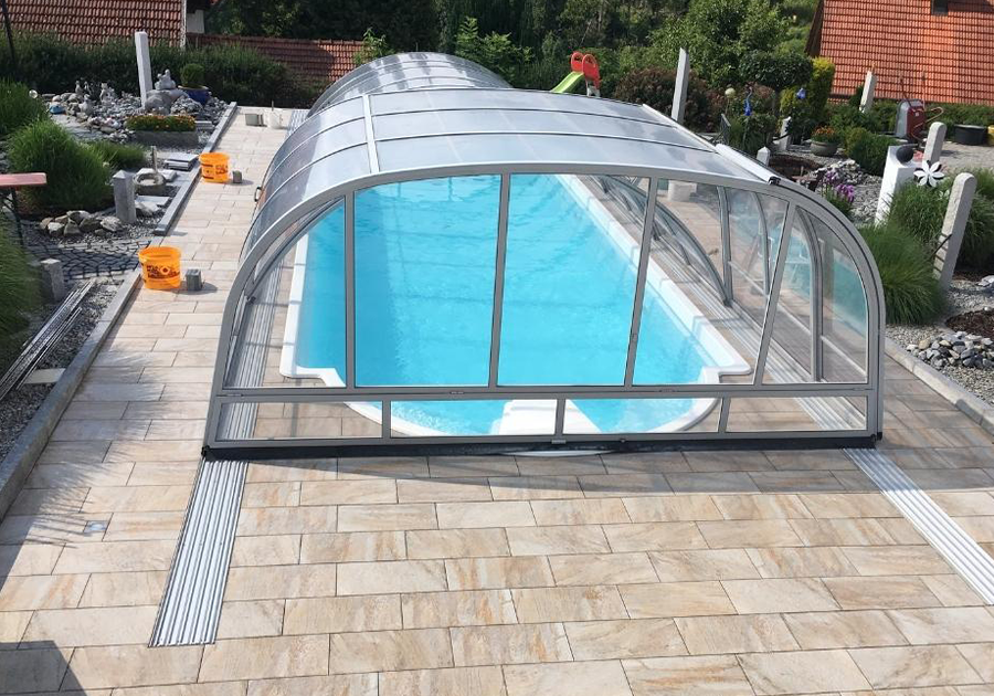 Das Bild zeigt einen rechteckigen Außenpool mit klarem, blauem Wasser, der von einer transparenten, gewölbten Überdachung aus Glas oder Kunststoff geschützt wird. Der Pool befindet sich auf einer hellen Stein- oder Fliesenfläche, die den gesamten Bereich umgibt. Im Hintergrund sind Gartenanlagen, Pflanzen und Häuser mit roten Dächern zu sehen.