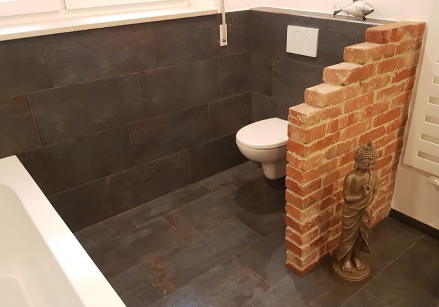 Modernes Badezimmer mit dunklen Fliesen. Im Vordergrund befindet sich eine weiße Toilette mit weißem Drücker. Das WC ist durch eine halbhohe Wand in Backsteinoptik abgetrennt. Davor steht eine kleine Dekorationsfigur auf dem Boden.