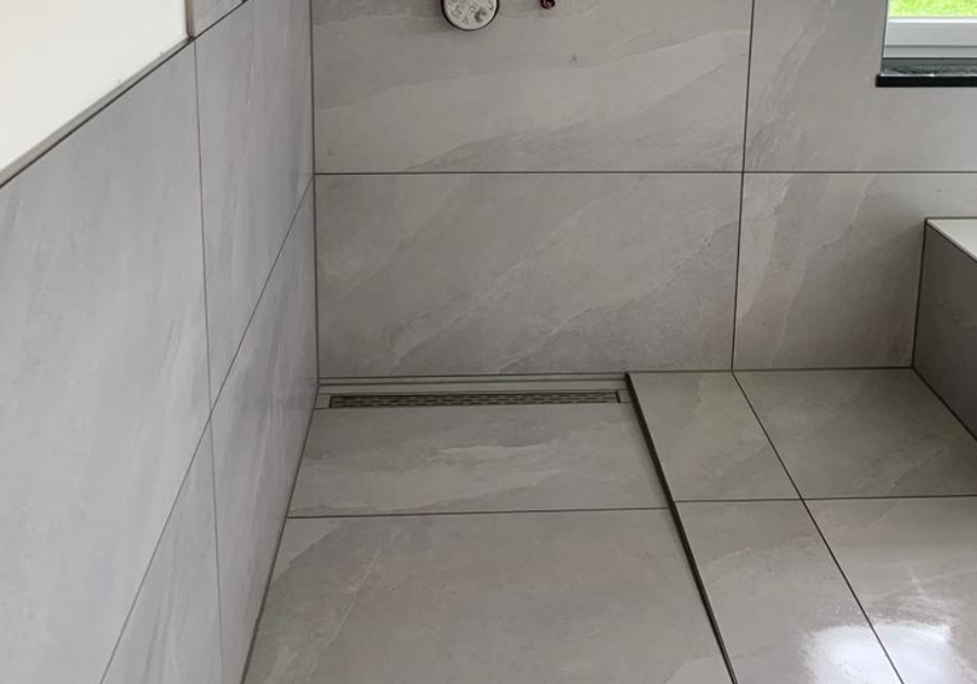 Bodengleiche Walk-in-Dusche mit großformatigen, hellgrauen Fliesen. Eine schmale Linienentwässerung (Duschrinne) ist bündig entlang der Wand eingelassen. An der Wand sind Wasseranschlüsse sichtbar, die Duscharmaturen werden noch installiert.