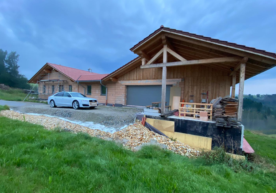 Das Bild zeigt einen Garagenanbau an einem Holzhaus mit rotem Ziegeldach. Vor der Garage liegt Baumaterial, und ein Auto ist auf der Zufahrt geparkt. Der Bereich um die Garage befindet sich noch im Bau, mit Kies, Holzstapeln und Werkzeugen im Vordergrund.